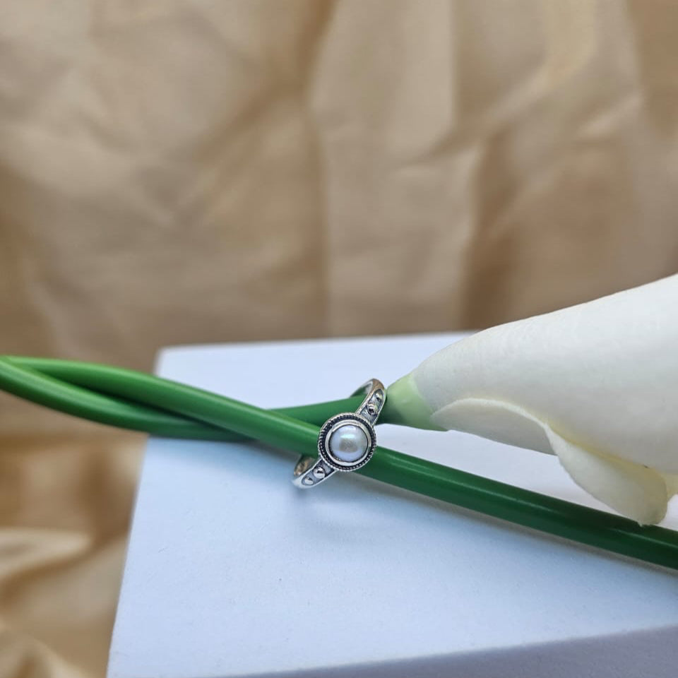 Silver ring with a pearl on a white flower and green stem against a beige background