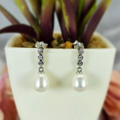 A pair of pearl earrings with a modern design, displayed in a white vase with a green plant background.