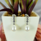 A pair of pearl earrings with a modern design, displayed in a white vase with a green plant background.