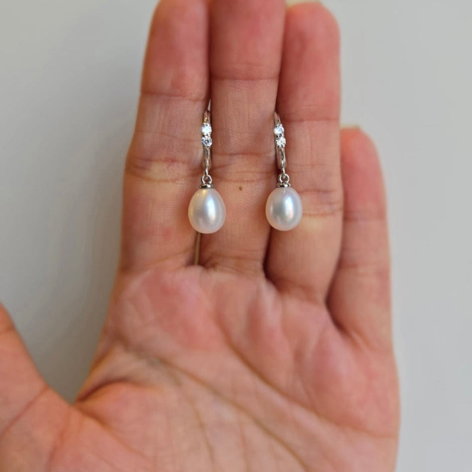 A pair of freshwater pearl drop earrings on sterling silver huggie hoops held in a person's hand.