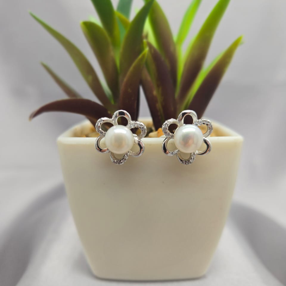 Pearl earrings with silver floral designs in front of a small potted plant on a light gray background
