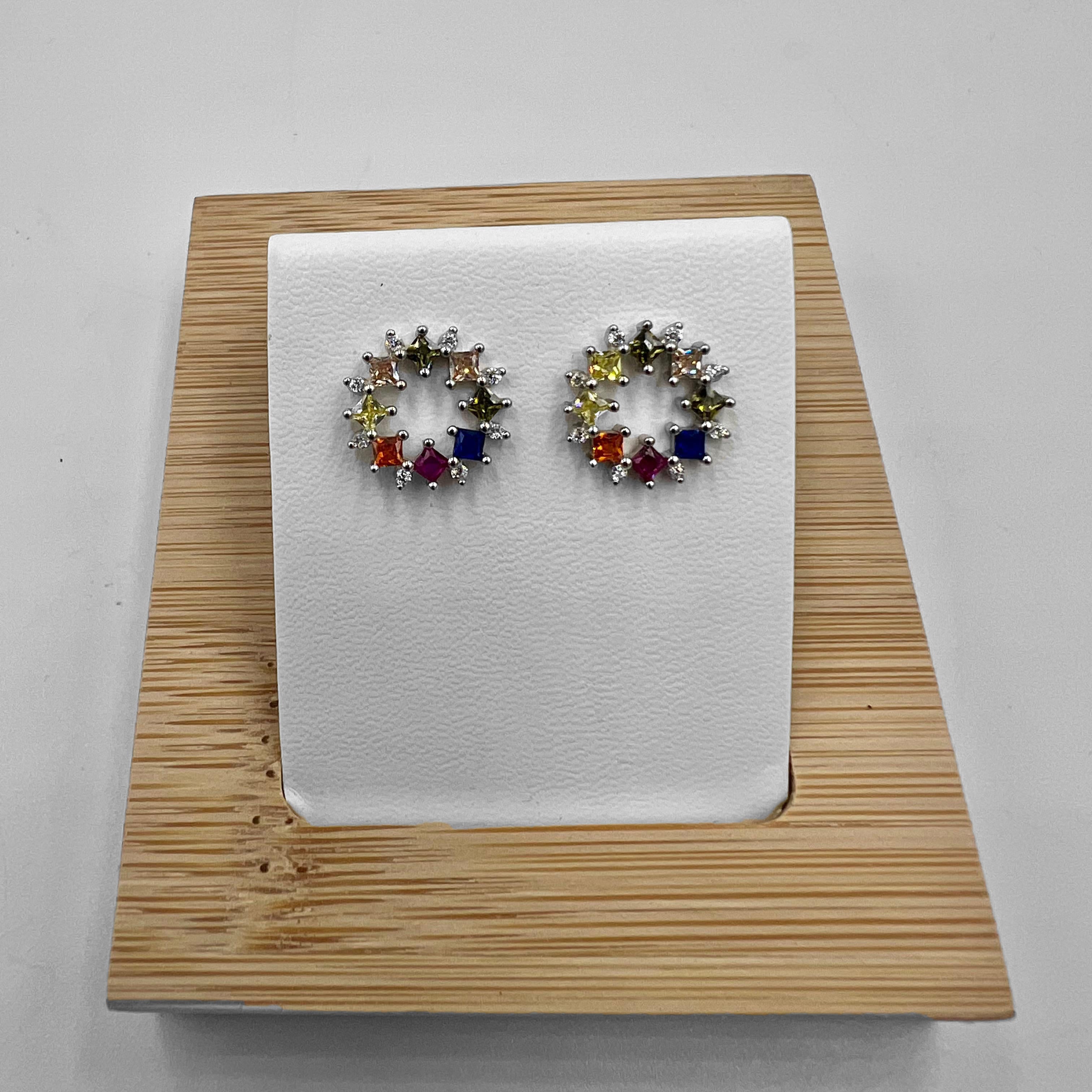 A pair of circle stud earrings crafted from sterling silver, adorned with colorful cubic zirconia stones, displayed on a white mount against a wooden background.