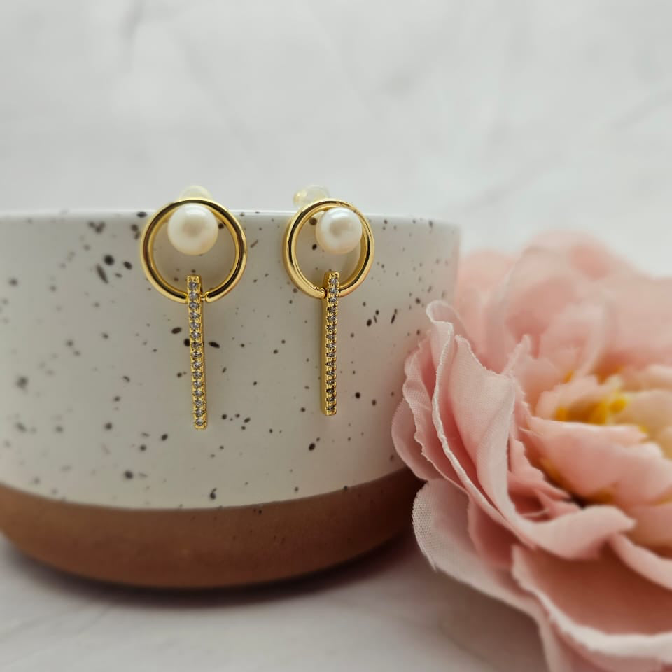 A pair of gold-plated drop earrings with pearl centers, displayed on a white surface next to a pink flower.