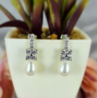 A pair of sterling silver earrings featuring a cubic zirconia flower design and freshwater pearl embellishments, displayed in a white vase with a green plant in the background.