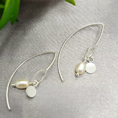 A pair of sterling silver drop earrings with pearl charms, displayed on a grey background with a green plant in the background.