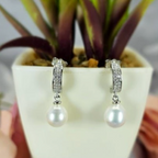 A pair of sterling silver drop earrings with freshwater pearl and cubic zirconia detail, displayed in a white vase with a green plant in the background.