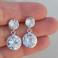 Silver earrings with clear gemstones held between fingers against a neutral background