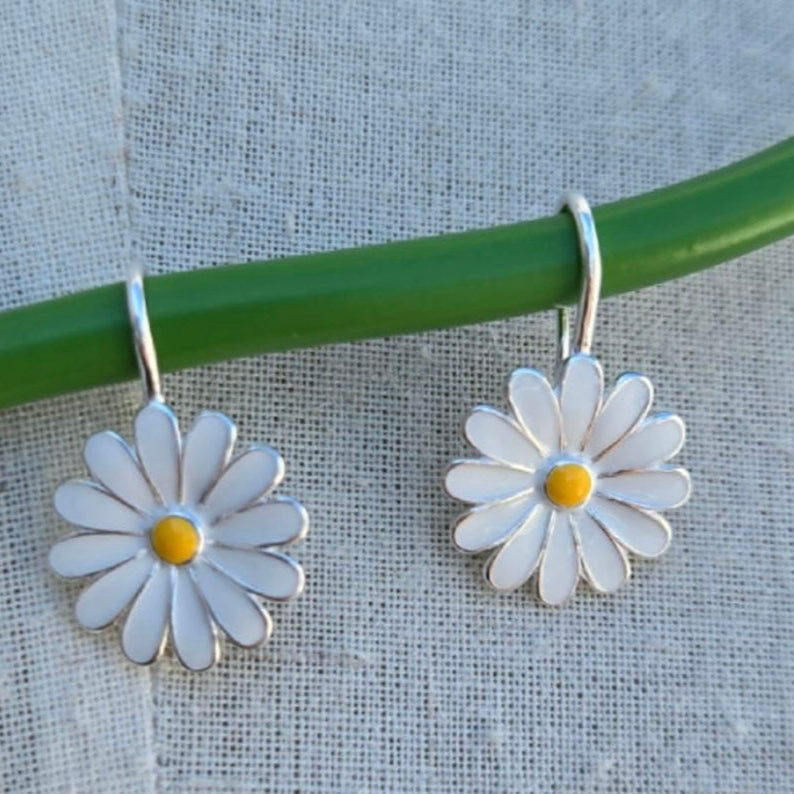 Daisy-shaped earrings hanging on a green rod against a textured background