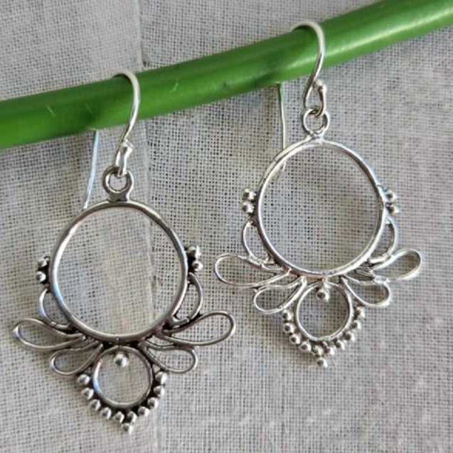 Silver hoop earrings with intricate designs hanging on a green stick against a gray background