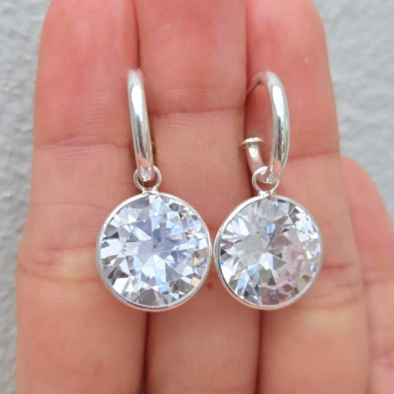 Silver earrings with clear gemstones held between fingers against a neutral background