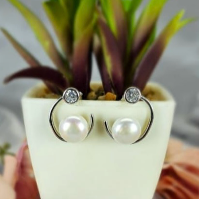A pair of modern design earrings featuring 17mm freshwater pearl and sparkling cubic zirconia, displayed in a white container with a green plant in the background.