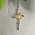 A sterling silver cross pendant with a rose gold inset, displayed against a green leafy background.