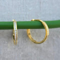 Gold hoop earrings with embedded stones on a green stick against a gray background