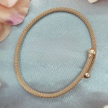 A rose gold plated sterling silver mesh fold over bracelet displayed on a grey surface with flowers in the background.