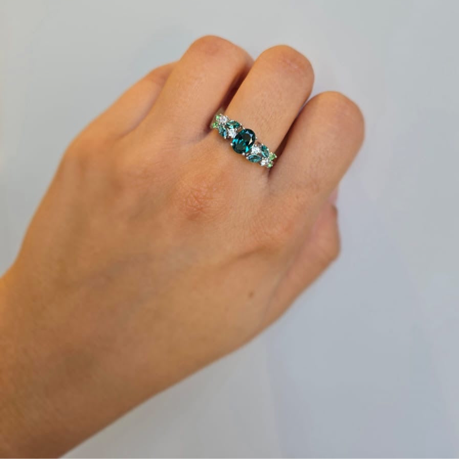 A hand wearing a green cubic zirconia cluster ring with white stones on a white background