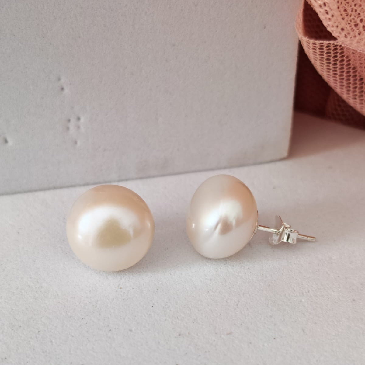 Pearl earrings on a light gray surface with a soft pink fabric background