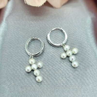 A pair of silver huggy earrings with freshwater pearl crosses on a grey fabric background with a floral accent.