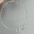 A sterling silver bracelet with three interlinked rings, displayed on a surface with a background of soft focus flowers.