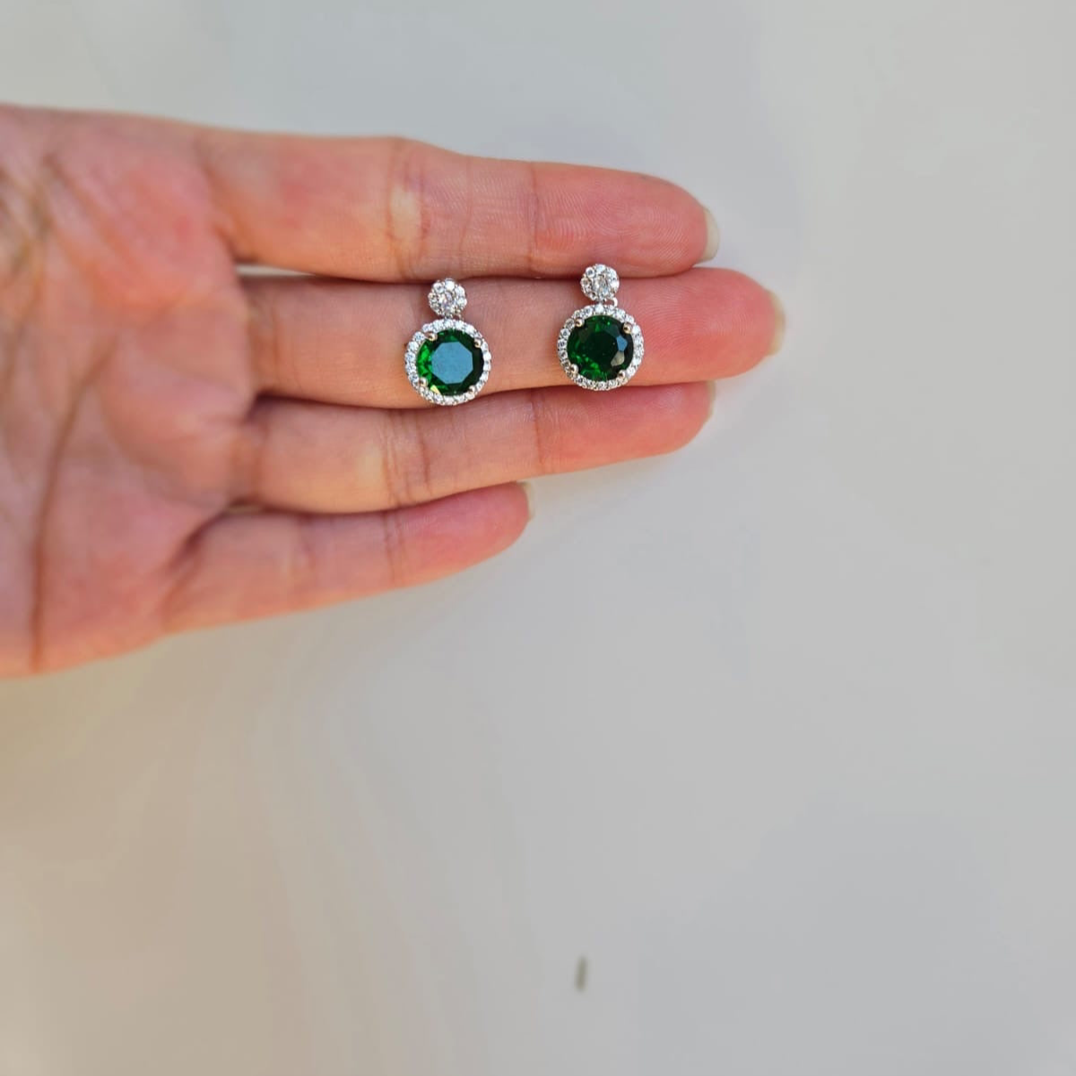 A pair of round green cubic zirconia stud earrings held in a person's hand, with a white background.