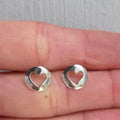 Silver heart-shaped earrings held between fingers on a neutral background