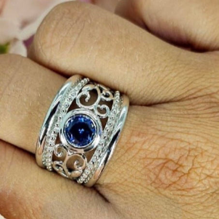 A sterling silver filigree detail ring with a blue cubic zirconia, held on a person's finger.