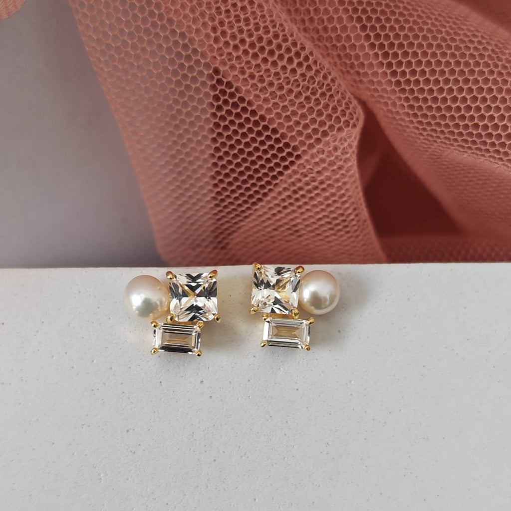 Pair of pearl and crystal stud earrings on a light gray surface with pink fabric in the background.