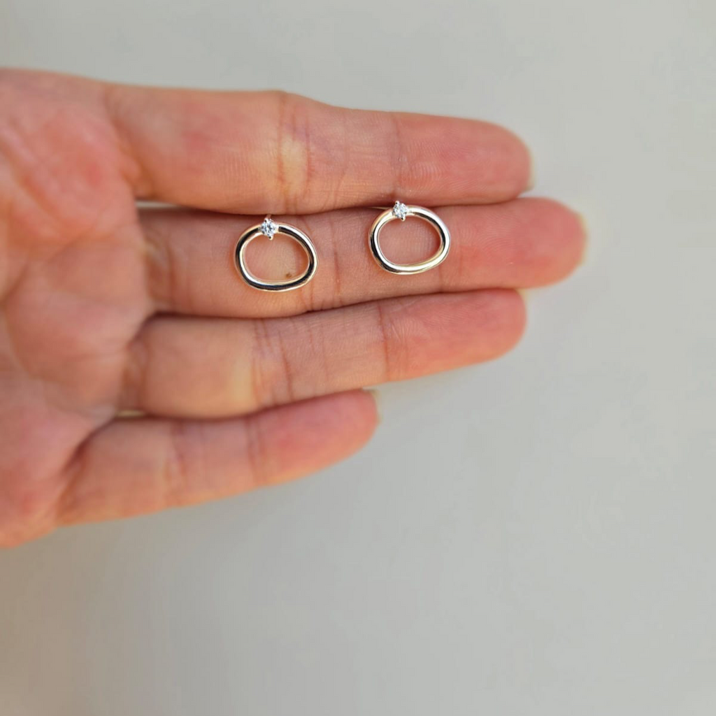 A pair of sterling silver open oval stud earrings with cubic zirconia, displayed on a hand.