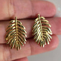 Gold leaf-shaped earrings held between fingers against a neutral background