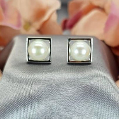 A pair of sterling silver square stud earrings with freshwater pearls on a grey fabric surface with a background of pink flowers.