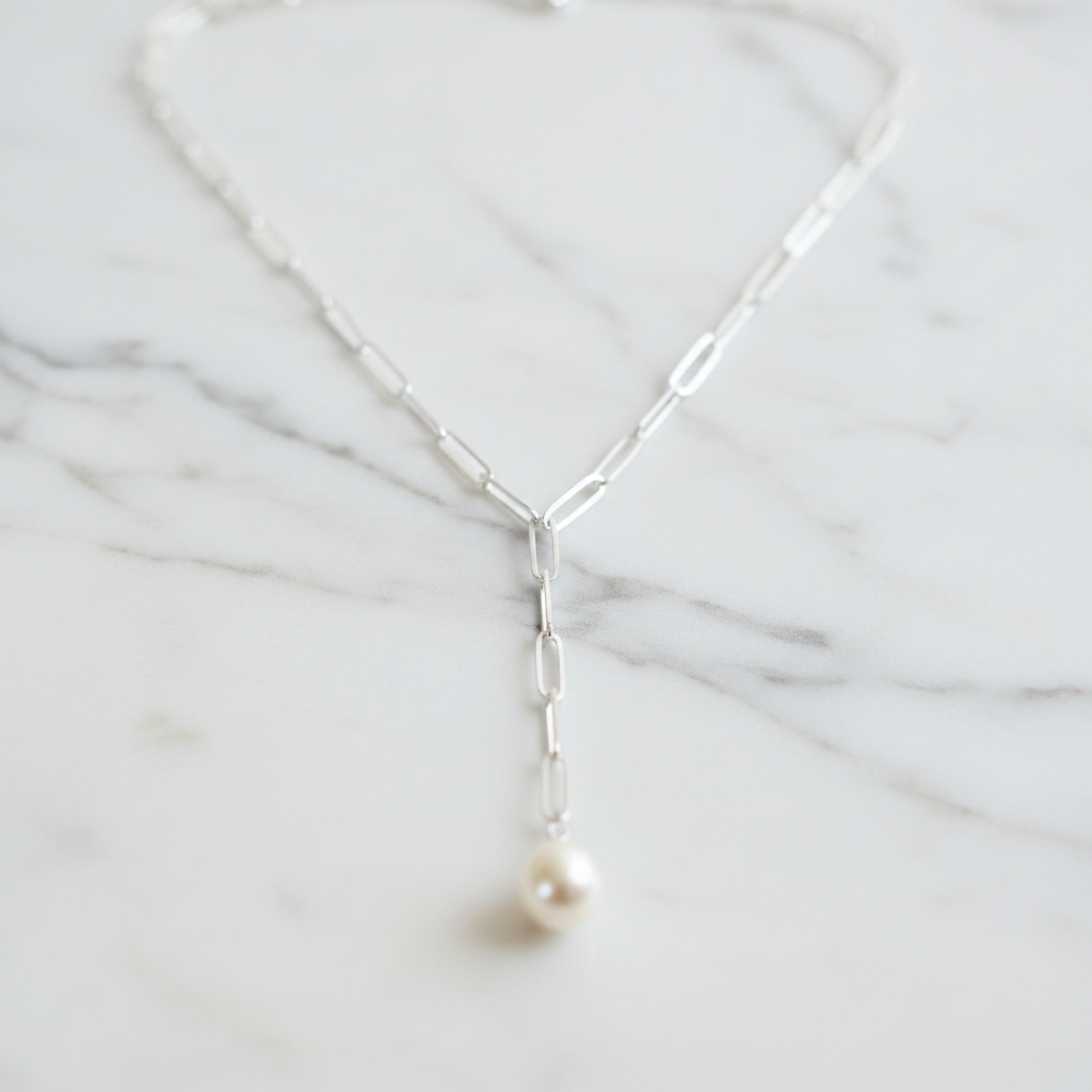 A sterling silver paperclip necklace with a freshwater pearl on one end, displayed on a wooden surface with flowers in the background.