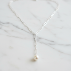 A sterling silver paperclip necklace with a freshwater pearl on one end, displayed on a wooden surface with flowers in the background.