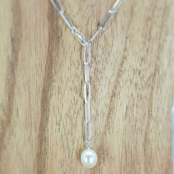 A sterling silver paperclip necklace with a freshwater pearl on one end, displayed on a wooden surface with flowers in the background.