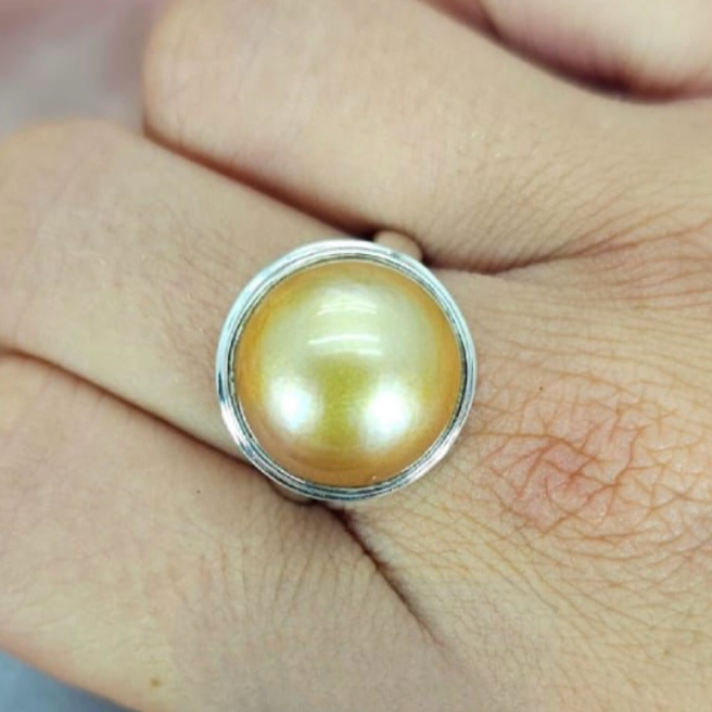 A gold/brown colored Mabe pearl ring set in a silver tube setting, displayed on a person's finger.
