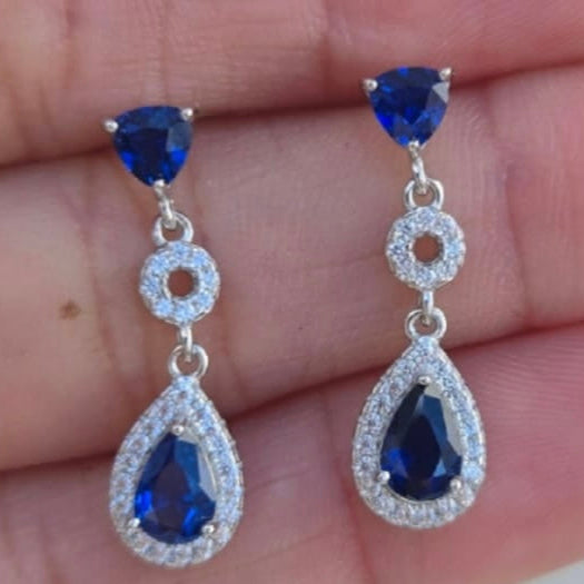 Sapphire teardrop earrings held between fingers against a blurred background