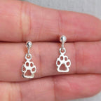 Silver paw print earrings held between fingers on a neutral background