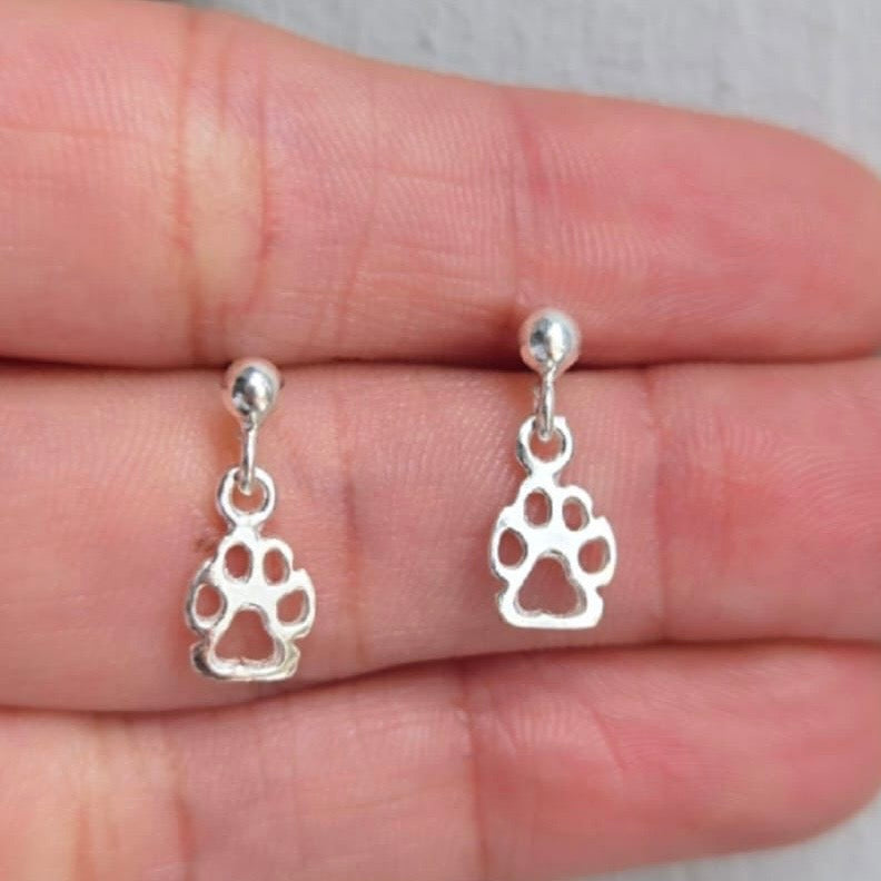 Silver paw print earrings held between fingers on a neutral background