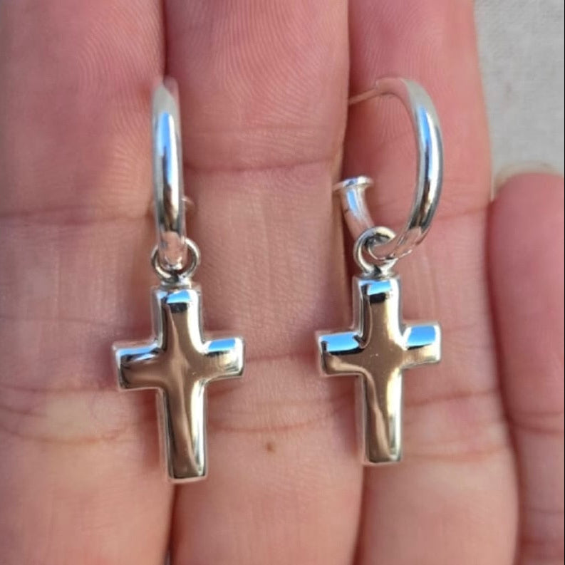 Silver cross earrings held on a hand against a neutral background