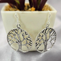A pair of silver drop earrings with a tree of life design, displayed in front of a potted plant.