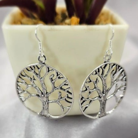 A pair of silver drop earrings with a tree of life design, displayed in front of a potted plant.