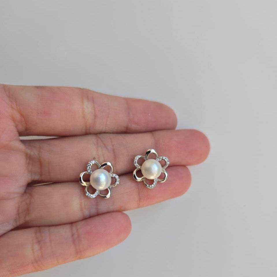 A pair of sterling silver flower pearl studs earrings held in a person's hand, displaying the earrings' floral design with a central pearl and small diamond-like accents.