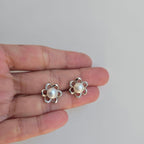 A pair of sterling silver flower pearl studs earrings held in a person's hand, displaying the earrings' floral design with a central pearl and small diamond-like accents.