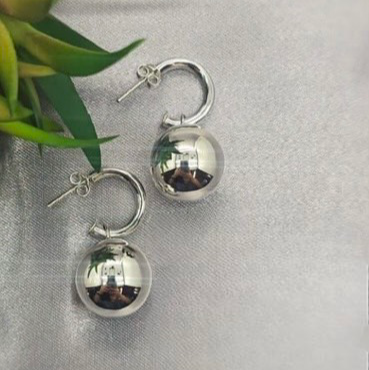 A pair of sterling silver earrings in a design that features a small spherical pendant attached to a larger hoop, displayed against a silver background with a green leaf.