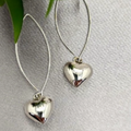 A pair of sterling silver heart drop earrings suspended by long wires against a grey background with green leaves partially visible to the side.
