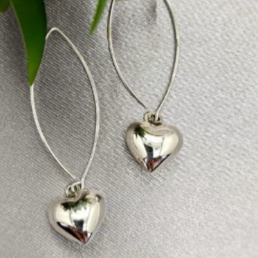 A pair of sterling silver heart drop earrings suspended by long wires against a grey background with green leaves partially visible to the side.