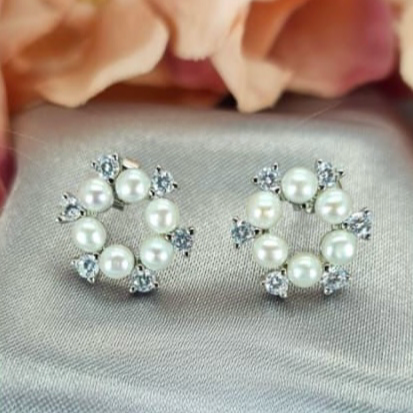 A pair of round 15mm freshwater pearl stud earrings with cubic zirconia stones in between, placed on a silky surface with a floral background.