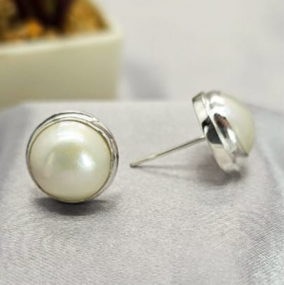 A pair of 14mm white mabe pearl stud earrings on a textured silver-colored surface.