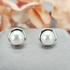 A pair of classic freshwater pearl stud earrings made of sterling silver, displayed against a floral background.