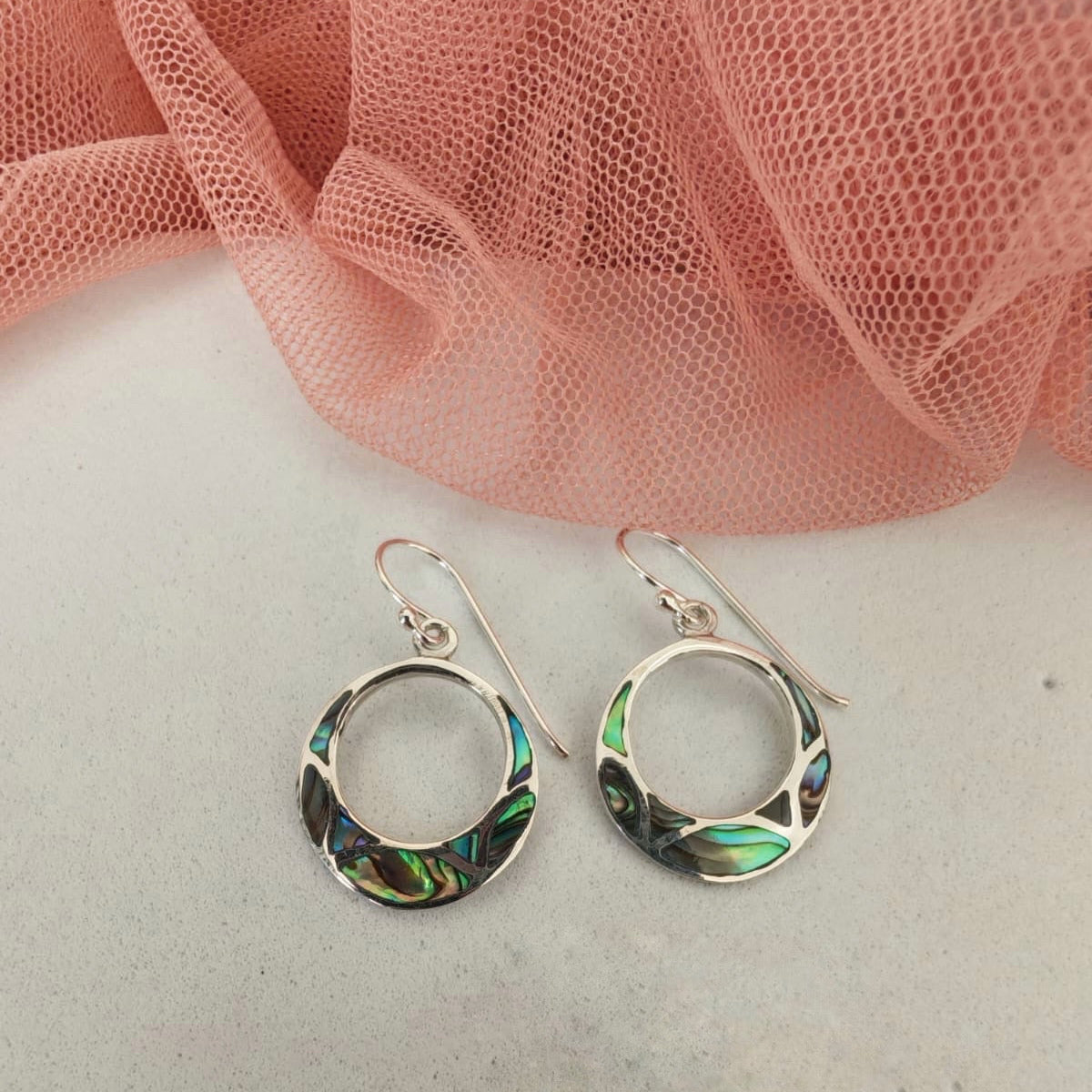 Silver earrings with abalone shell inlay on a white surface with pink fabric in the background