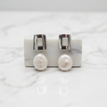 Pearl earrings with silver hooks displayed in front of a small potted plant on a gray background