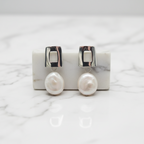 Pearl earrings with silver hooks displayed in front of a small potted plant on a gray background